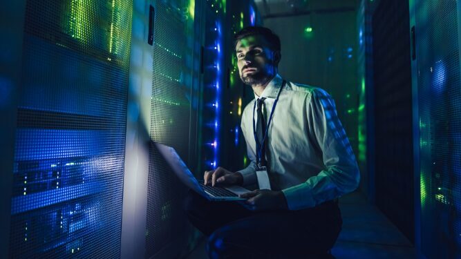 man is working on data centre health and safety on a laptop in a dimly lit network server room