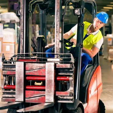 forklift driver wearing ppe driving a forklift in a warehouse leans out to the side from his driving position, highlighting the importance of a warehouse safety audit in identifying things that are often overlooked, such as potential safety incidents like falling from a forklift
