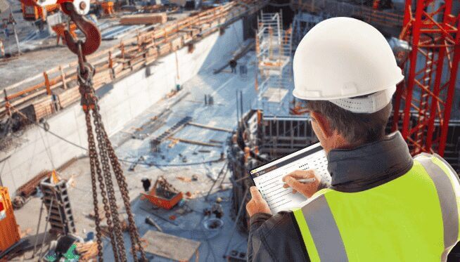 Aerial view of a construction site behind a man holding a tablet, using it to implement risk management services.