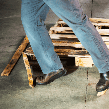 Near miss reporting example: Worker's boot catches on broken wooden pallet and debris on a warehouse floor, illustrating a tripping hazard and potential safety incident.