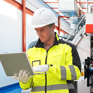 A construction or factory worker in a white hard hat, high-visibility yellow and black jacket, and gloves is intently looking at and typing on a silver laptop. The background shows industrial equipment and a clean manufacturing/processing environment, suggesting he is using the laptop for on-site risk assessment or management with risk register software.