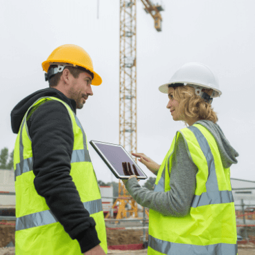 Two construction workers, one male in a yellow hard hat and one female in a white hard hat, both wearing high-visibility vests, stand on a construction site. The female worker is pointing at a tablet she is holding while talking to the male worker. In the background is a large crane. This image illustrates a site-specific induction being conducted.