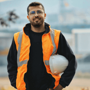 A smiling male construction worker wearing an orange high-visibility vest and safety glasses, holding a white hard hat. The image represents the smooth on-boarding process with contractor induction software.